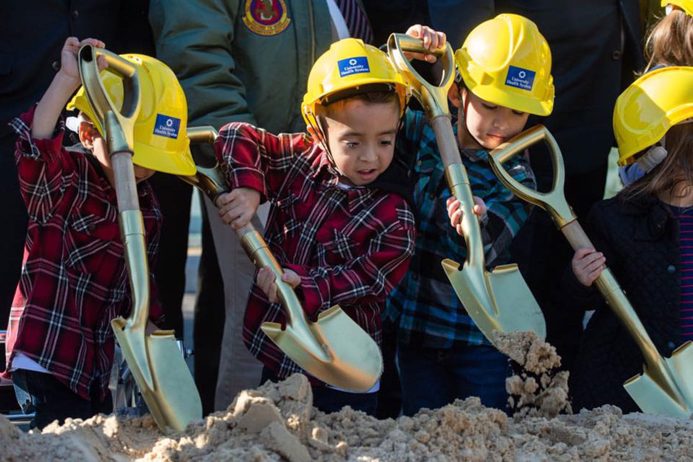 Kids at a hospital groundbreaking event digging dirt with a shovel and a yellow hardhat.