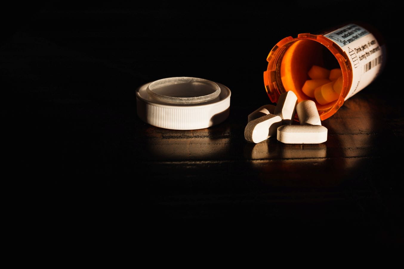 Pills spill out of an open pill bottle laying on its side in the dark.