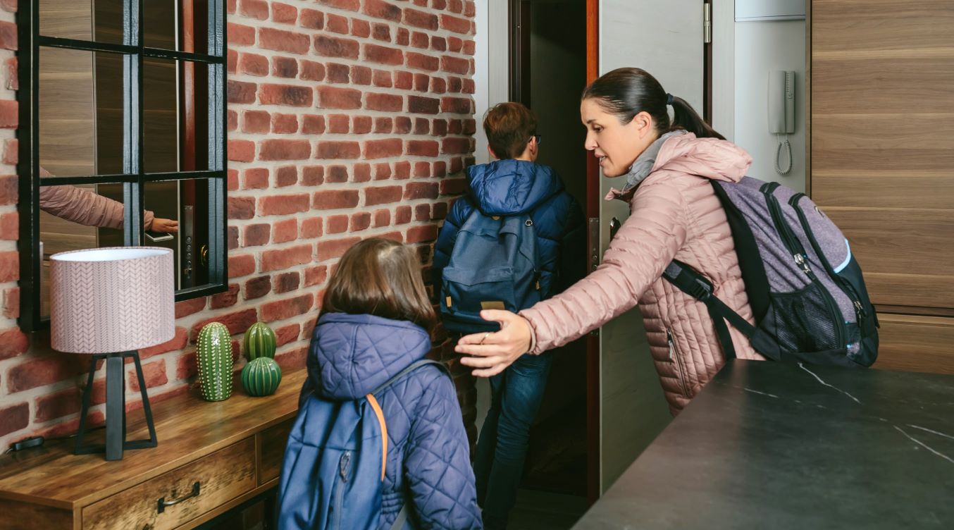 A woman guides two children wearing backpacks out the front door.