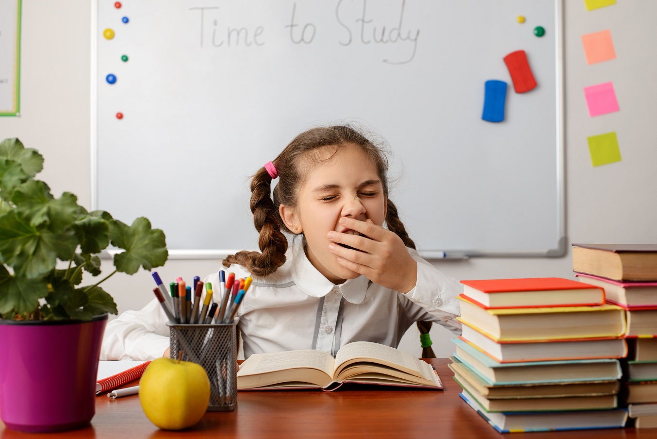 A young girl covers her mouth while she yawns, sitting at a desk in school