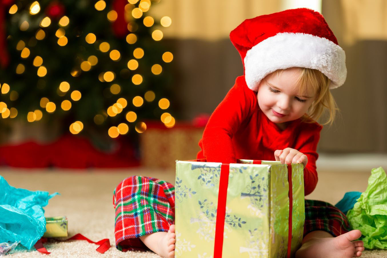 A little girl sits under a Christmas tree wearing a Santa hat. She's reaching into a gift box to pull out a toy.
