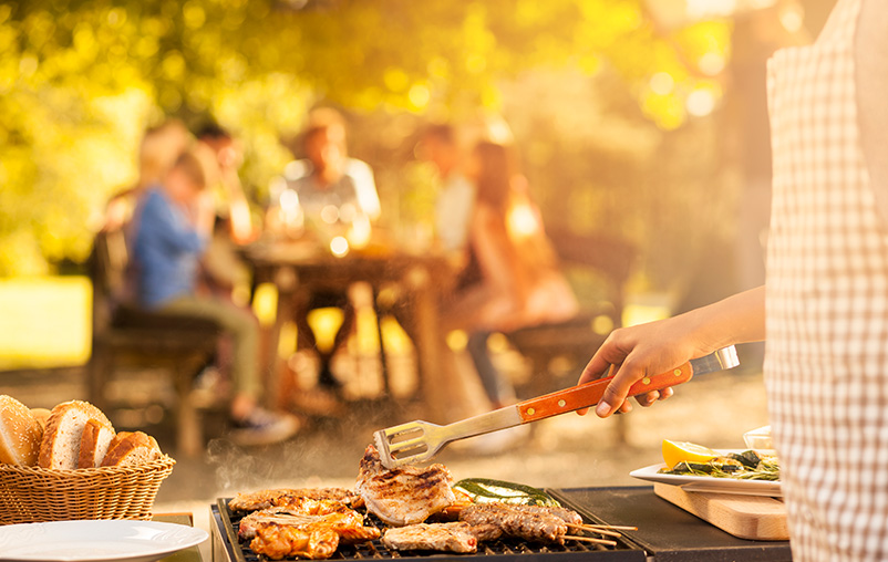 Close-up of a woman grilling on a barbecue with people sitting at a picnic table in the background.