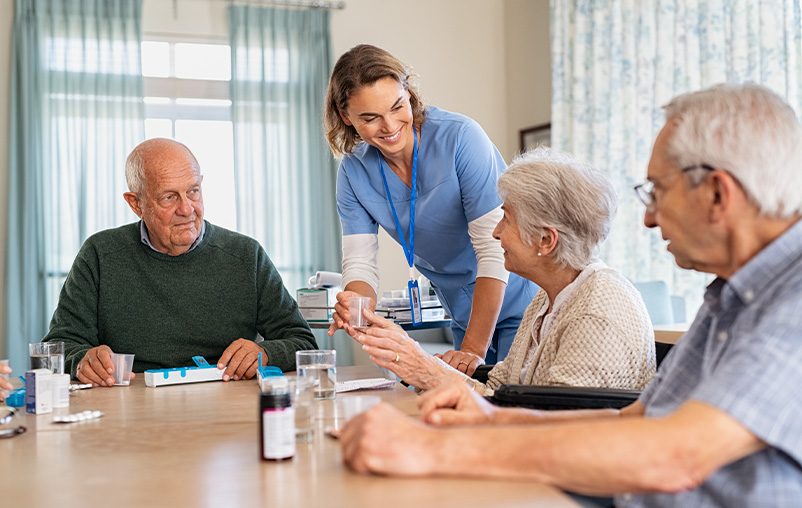 Three elderly people sit around a table and a nurse hands one of them a cup of water.
