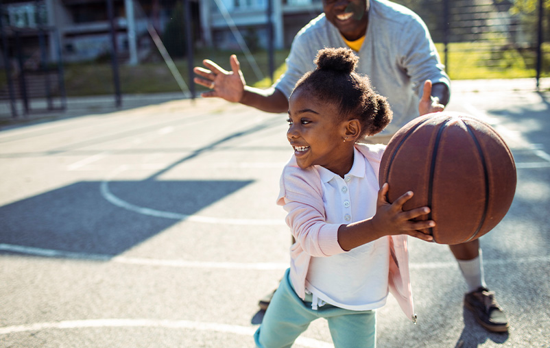 A smiling little girl plays basketball. A smiling man, maybe her dad, stands behind her.