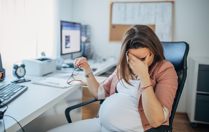 A pregnant woman sits at her desk and has her head in her hand; she looks stressed.