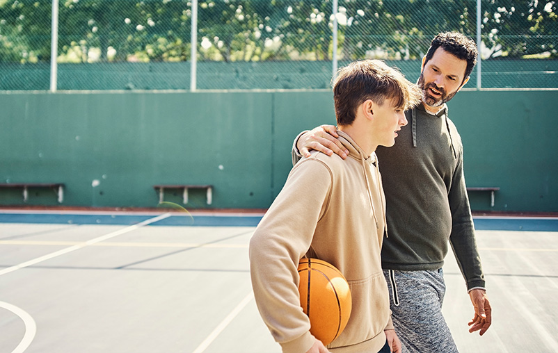 A dad and teenage son talk on a basketball court. The dad has his arm around his son's shoulders.