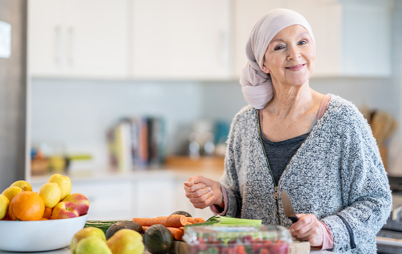 A woman wearing a headscarf stands by the kitchen counter, which has a colourful array of vegetables on it. She smiles as she chops the produce.