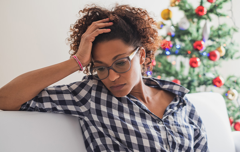 A woman looks stressed out sitting in front of a Christmas tree indoors.