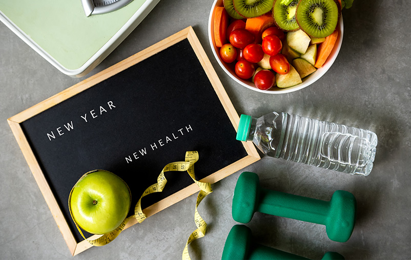 "New Year New Health" spelled on a sign, bowl of fruit, weights, water bottle.