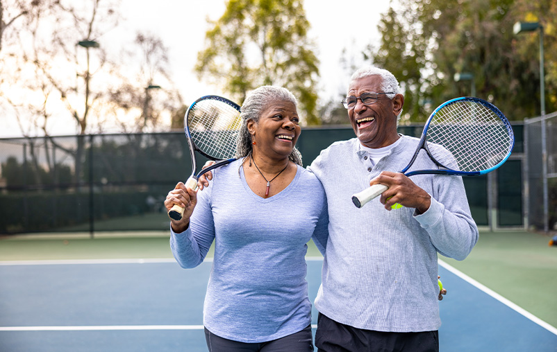 A man and woman smile, holding tennis racquets on a tennis court.