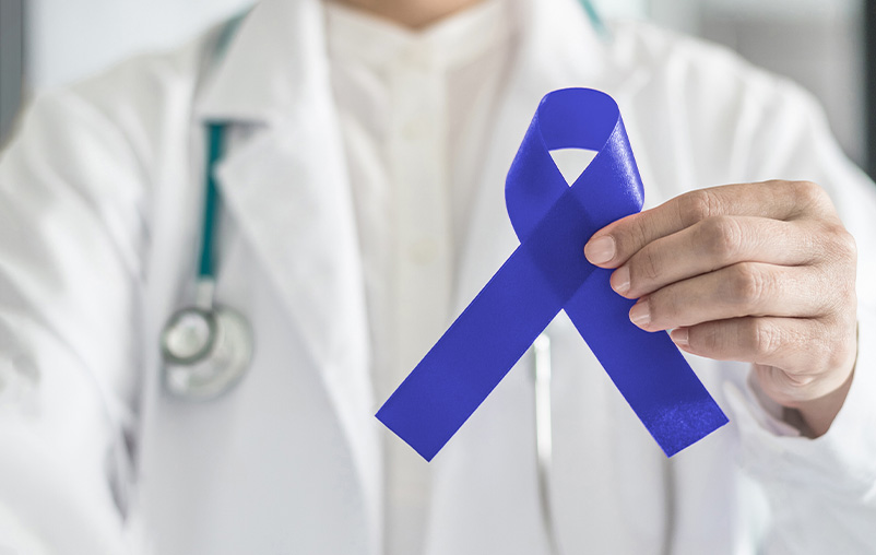 A person in a white lab coat holds a blue ribbon to represent colorectal cancer awareness.
