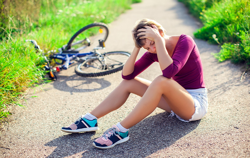 A woman sits on the ground and holds her head in pain. A crashed bike is in the background.