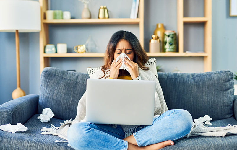 A woman sitting on a couch in her home sneezing into a tissue while on a laptop.