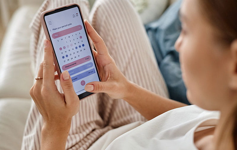 Young adult woman holding smartphone while checking menstrual cycle calendar app.