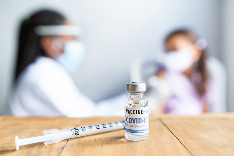COVID-19 booster shot in focus with a child receiving the vaccine blurred in the background.
