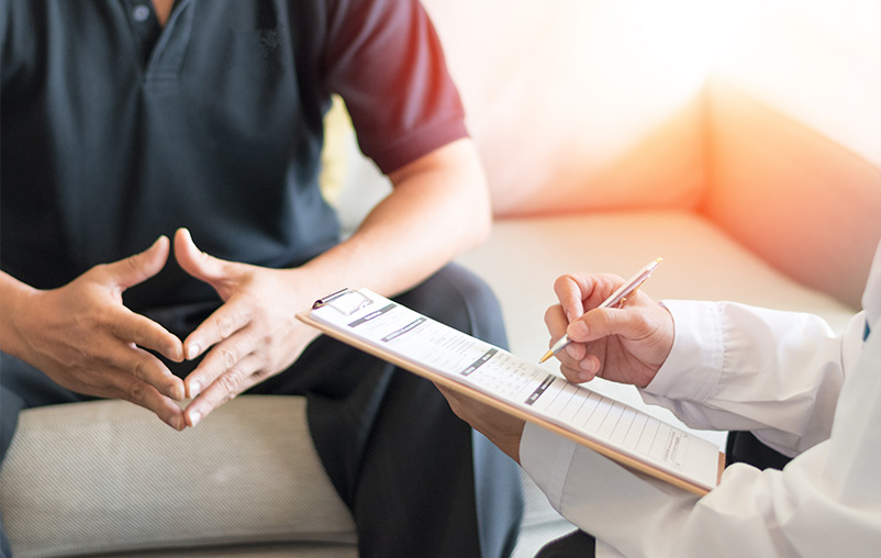 A man sits and talks with a health care provider, who is holding a clipboard.