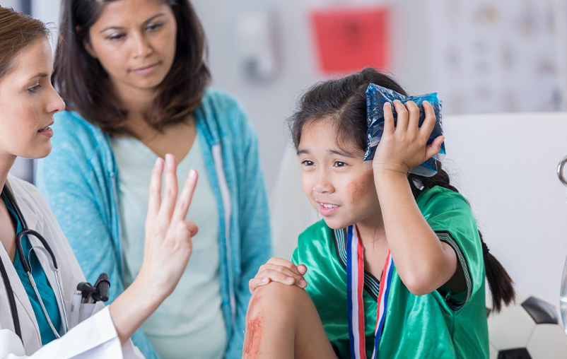 A young girl in a soccer uniform holds an ice pack to her head while a medical provider evaluates her for a concussion.