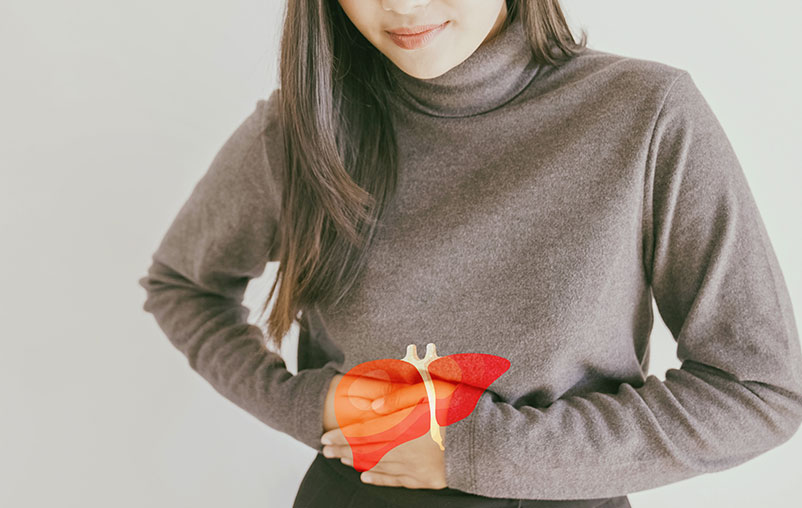 Woman in a gray sweater and black dress pants holding her hands above the upper right quadrant of her abdomen where her liver is located with a small digital graphic of a liver with subtle transparency over her hands.