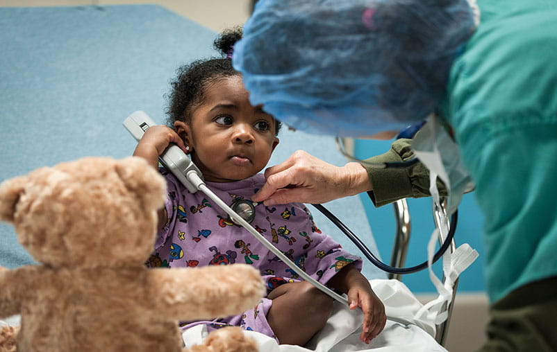 A young child in the Congenital Heart program sits on a bed as a University Health provider listens to her heart.
